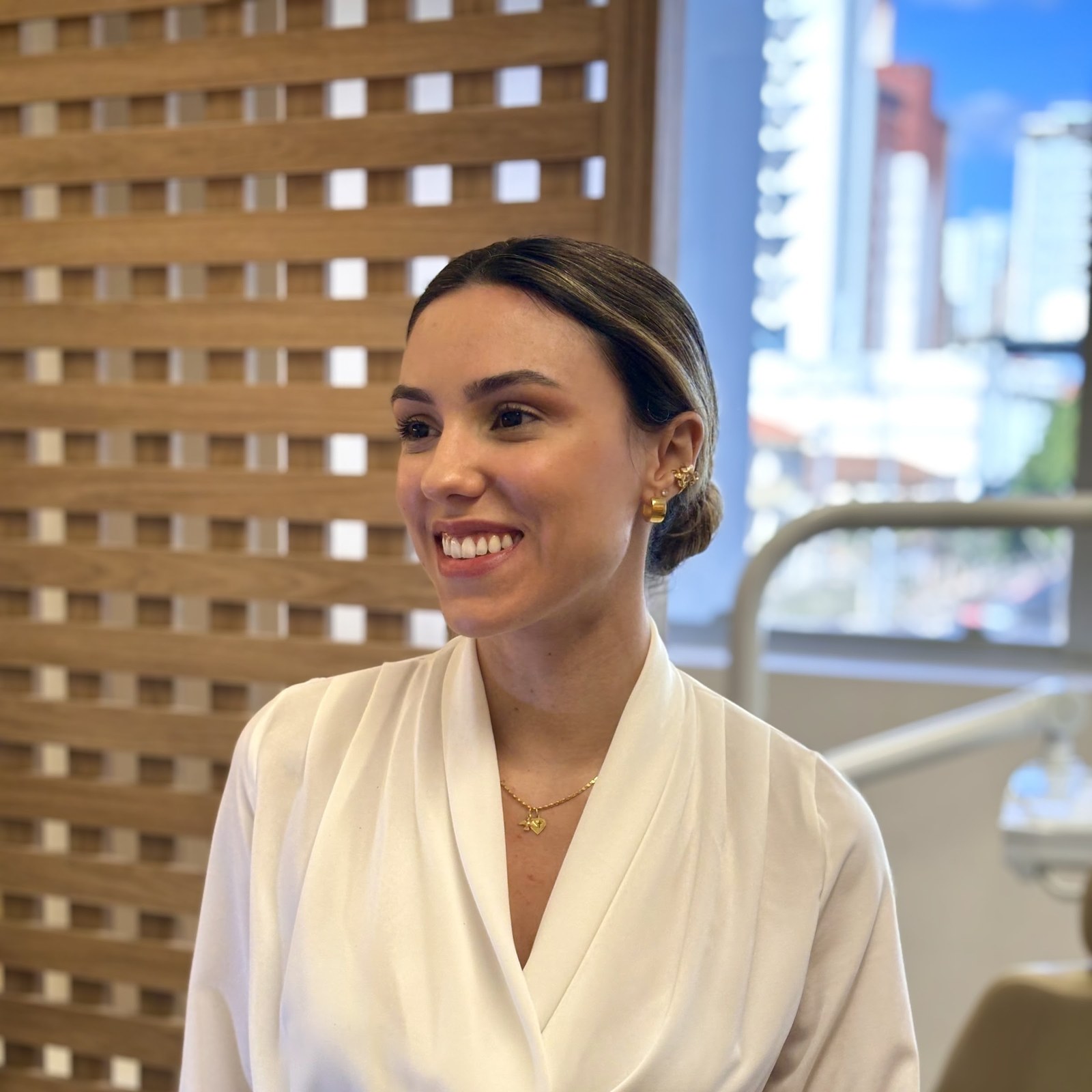 Dra. Clara Braga em retrato no consultório do Belvedere, blusa creme e coque baixo, com painel ripado de madeira ao fundo